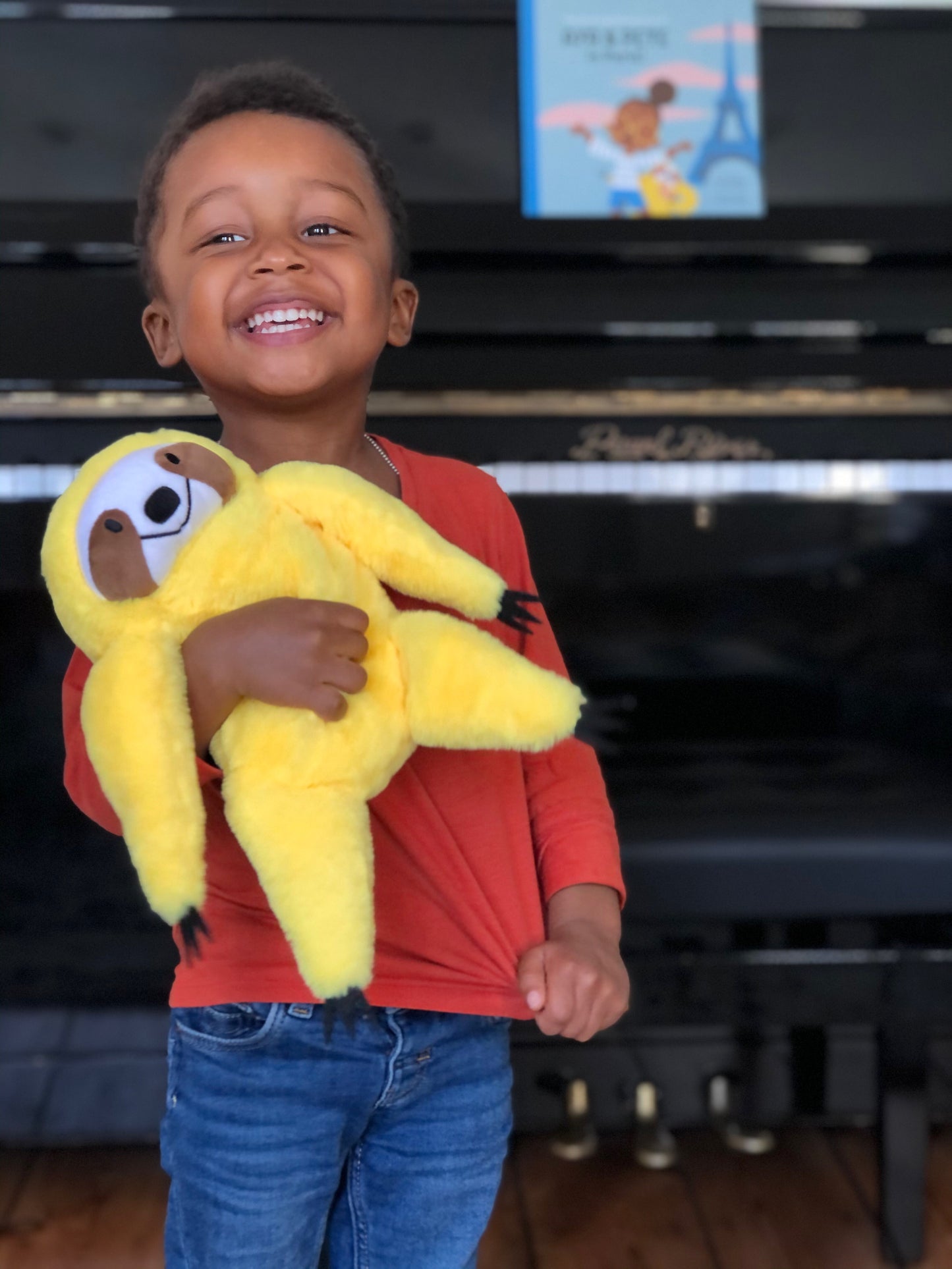 Little boy in a red shirt smiling and holding a yellow plush sloth, Pete. A piano and Aya & Pete in Paris appears in the background.