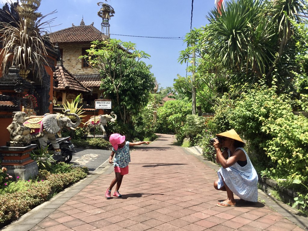 Image shows a young child in a pink hat posing for a photo with a woman stooped with a camera and a traditional hat on a leafy street. 